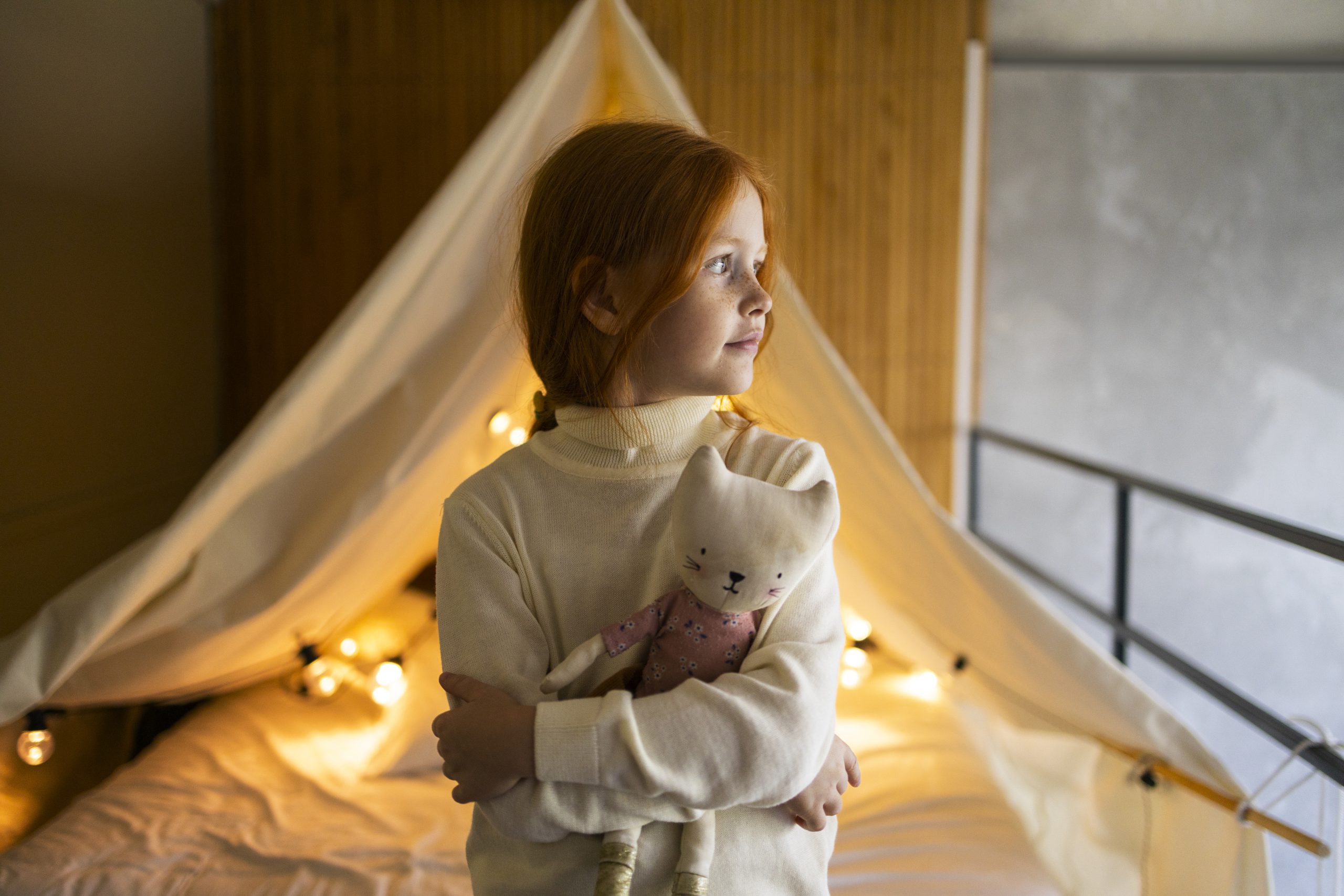 Dziewczynka trzymająca maskotkę przy łóżeczku domek dla dzieci oświetlonym lampkami